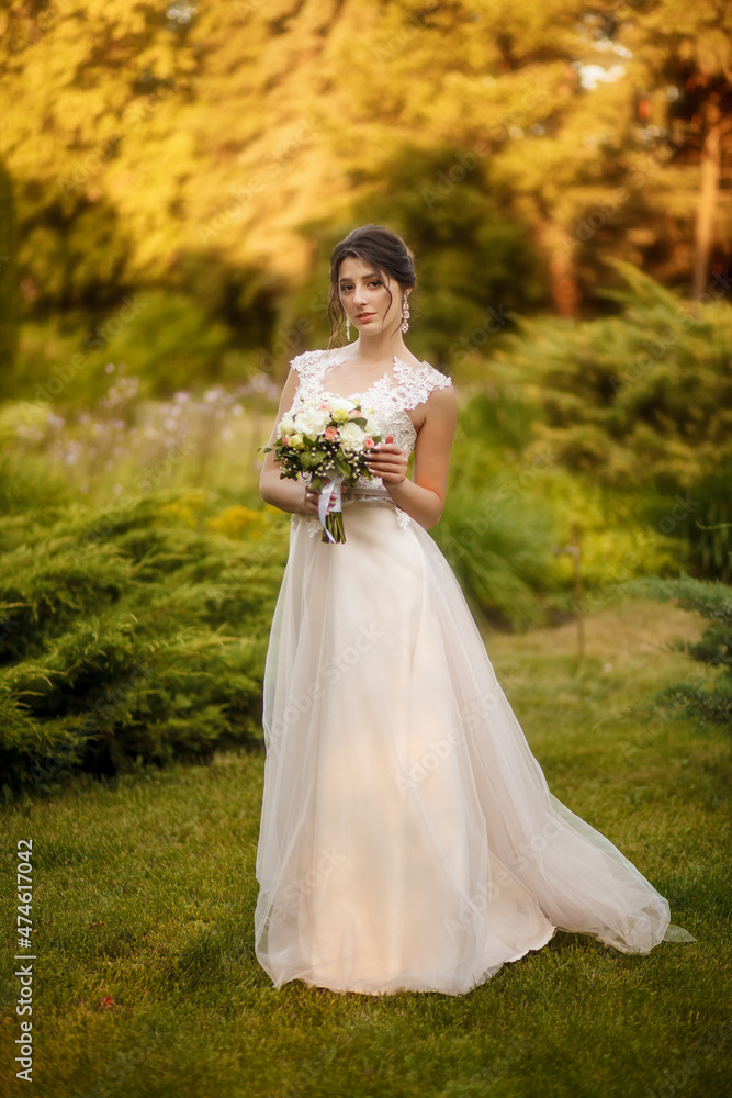 A beautiful bride in a white dress holds a wedding bouquet in her hand. Bride on a walk on a warm autumn day. Portrait of a happy wedding bride on their wedding day.