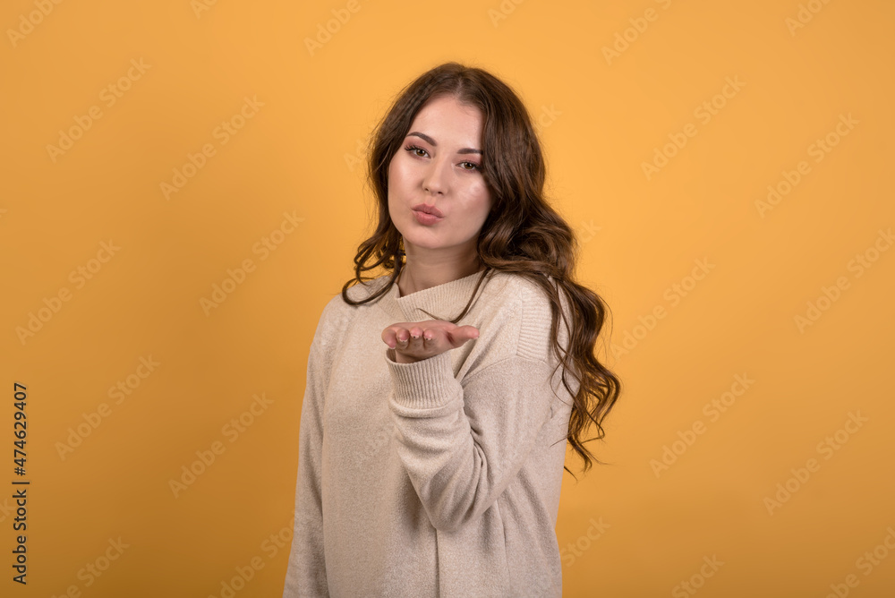 Attractive and happy caucasian brunette girl with wavy hair in a casual jumper showing a flying kiss on an orange studio background.