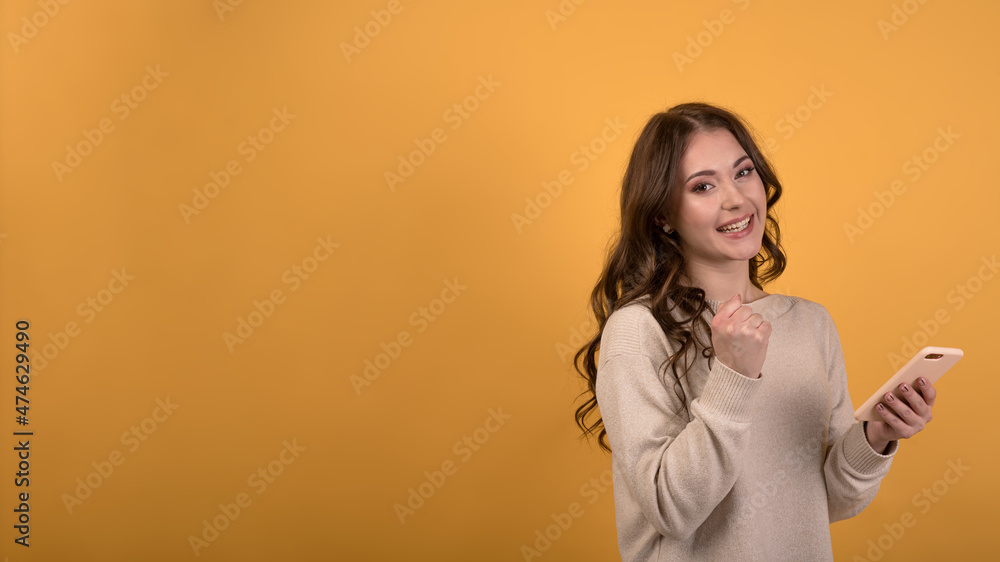 A young Caucasian brunette girl with wavy hair in a casual jumper holding a smartphone, looking at camera and enjoying victory on an orange studio background with copy space for advertising. A young Caucasian brunette girl with wavy hair in a casual jumper holding a smartphone, looking at camera and enjoying victory on an orange studio background with copy space for advertising.