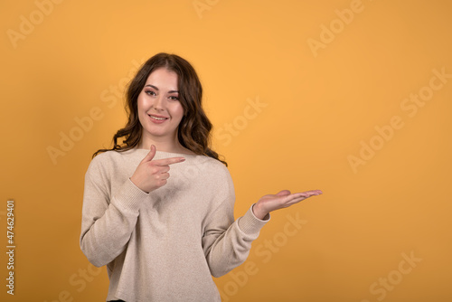 Attractive and happy Caucasian brunette girl with wavy hair in a casual jumper showing with her hand and pointing her finger at an empty space on the palm for copy space on an orange stage background. © Павел Костенко
