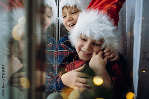 cute caucasian children wearing snta hats and plaid shirts sitting hugging by the window waitinf for christmas photo