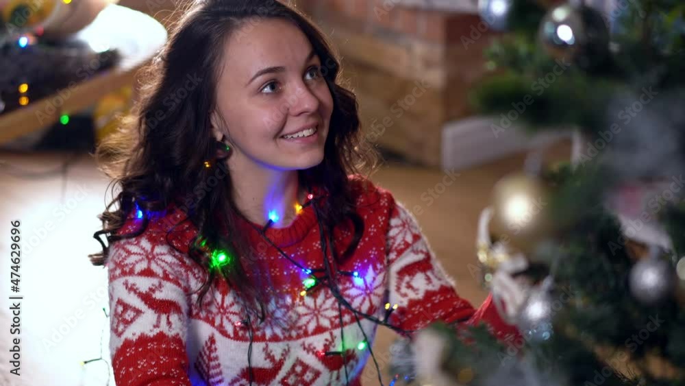 Joyful young slim woman singing decorating New Year tree with Christmas lights indoors. Portrait of excited happy Caucasian millennial lady enjoying holidays at home in living room Joyful young slim woman singing decorating New Year tree with Christmas lights indoors. Portrait of excited happy Caucasian millennial lady enjoying holidays at home in living room
