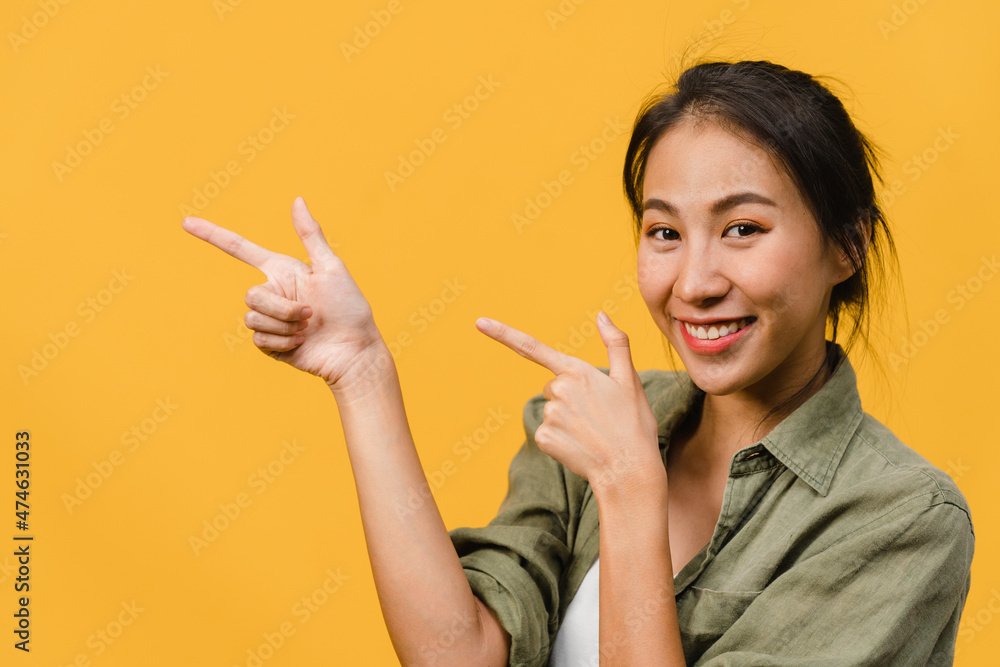 Portrait of young Asian lady smiling with cheerful expression, shows something amazing at blank space in casual cloth and looking at camera isolated over yellow background. Facial expression concept. Portrait of young Asian lady smiling with cheerful expression, shows something amazing at blank space in casual cloth and looking at camera isolated over yellow background. Facial expression concept.