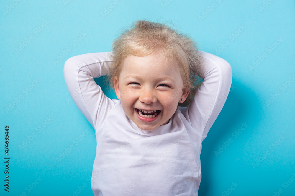 Naklejka premium Smiling beautiful blonde girl lying down holds her hands behind her head on a blue background. Top view, flat lay.