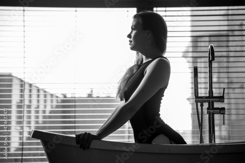 A young, slender girl in the bathroom stands naked against the background of a window with blinds.