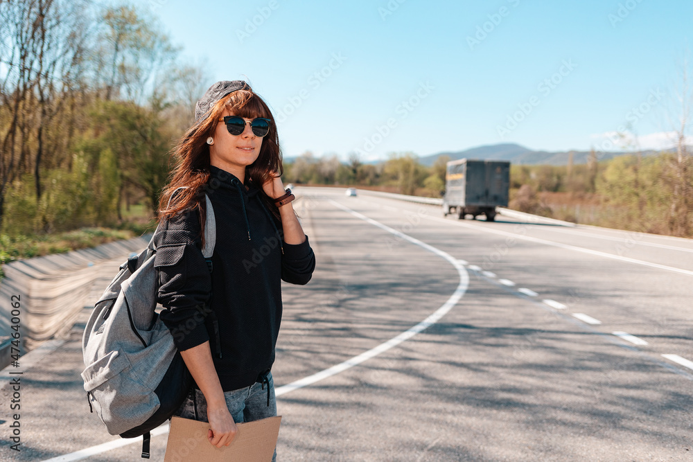 Fototapeta premium Hitchhiking. Smiling young woman in sunglasses and cap holding a cardboard sign. Country road on the background. Copy space. The concept of local traveling