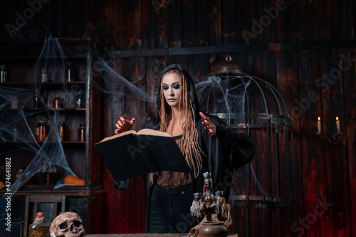 Halloween concept. Witch dressed black hood with dreadlocks standing dark dungeon room use magic book for conjuring magic spell