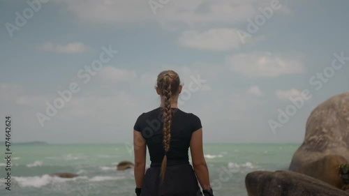 zoom out of athletically built female standing on the big stone by the ocean and looks ahead  photo