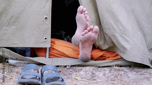 Bare Feet Male Legs Peeking Out from an Open Military Tent in Nature. Tourist is resting, sleeping at camping, airing naked legs. Traveler relaxes in a tent without slippers among trees outdoors.