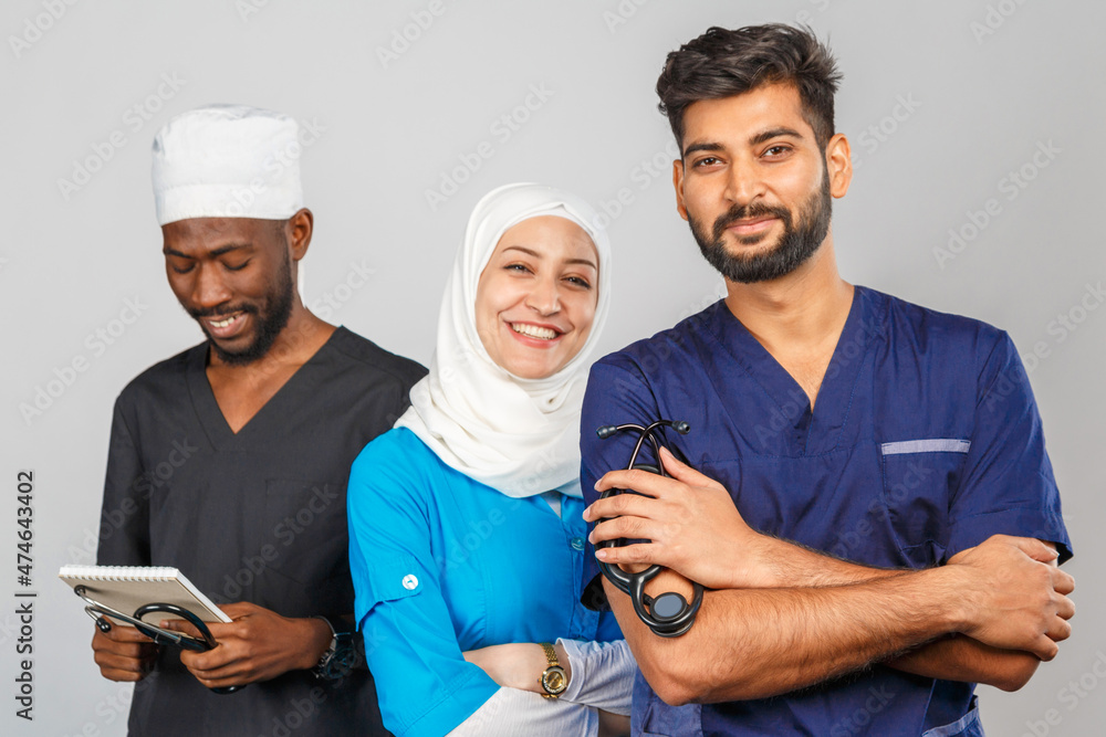 Fototapeta premium Portrait of positive healthcare workers standing in the hallway and looking at the camera. Group of medics smiling over gray background. muslim doctor woman on background of african and indian doctor