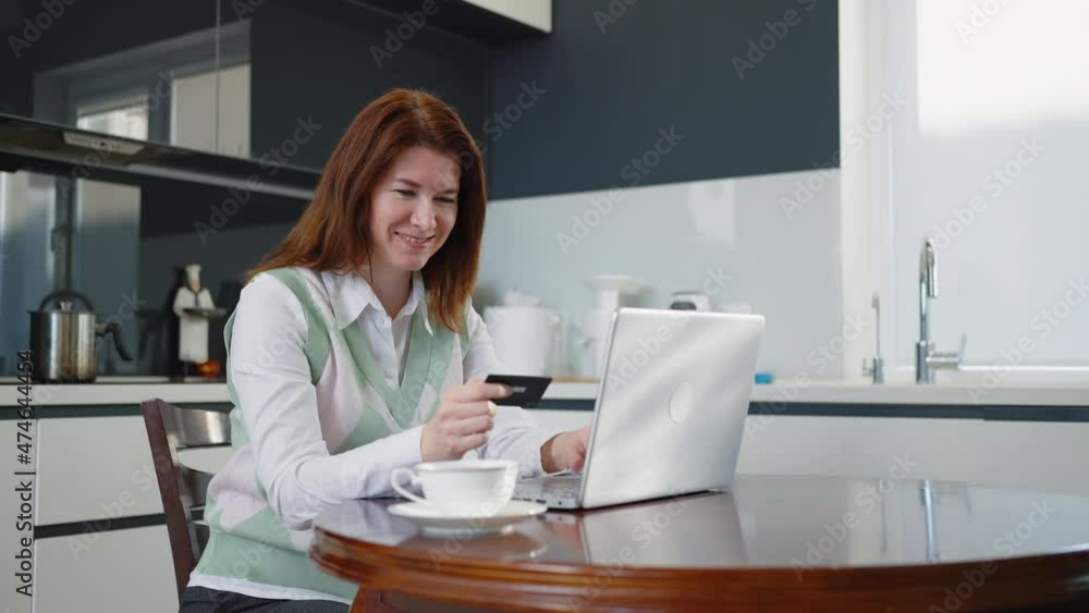 Smiling woman holding credit card and entering numbers on laptop, making online order while sitting in kitchen, making yes gesture and thumb up. Concept of online shopping Smiling woman holding credit card and entering numbers on laptop, making online order while sitting in kitchen, making yes gesture and thumb up. Concept of online shopping
