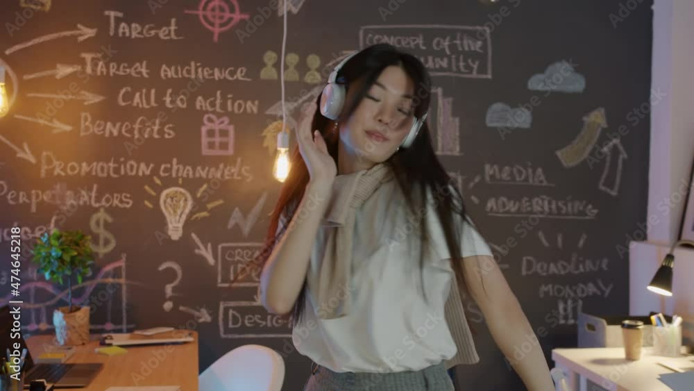 Beautiful Asian girl employee is dancing in creative workplace wearing headphones enjoying music in evening. Youth lifestyle and job concept. Beautiful Asian girl employee is dancing in creative workplace wearing headphones enjoying music in evening. Youth lifestyle and job concept.