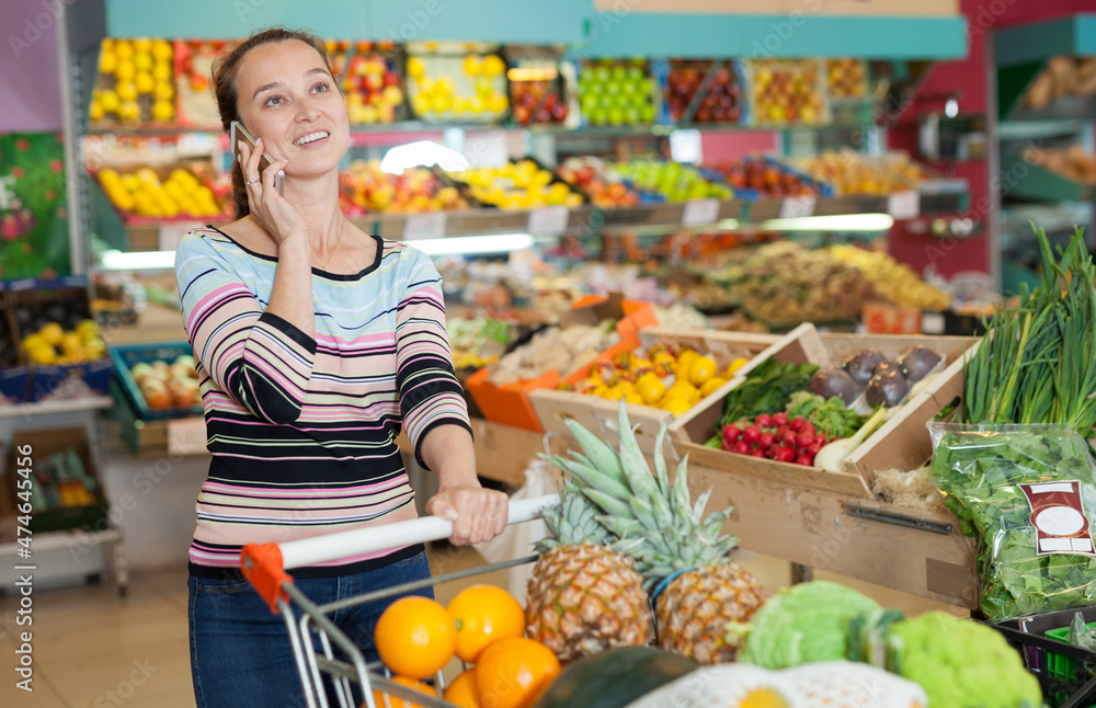Portrait of cheerful woman with shopping cart full of fruits and vegetables at store