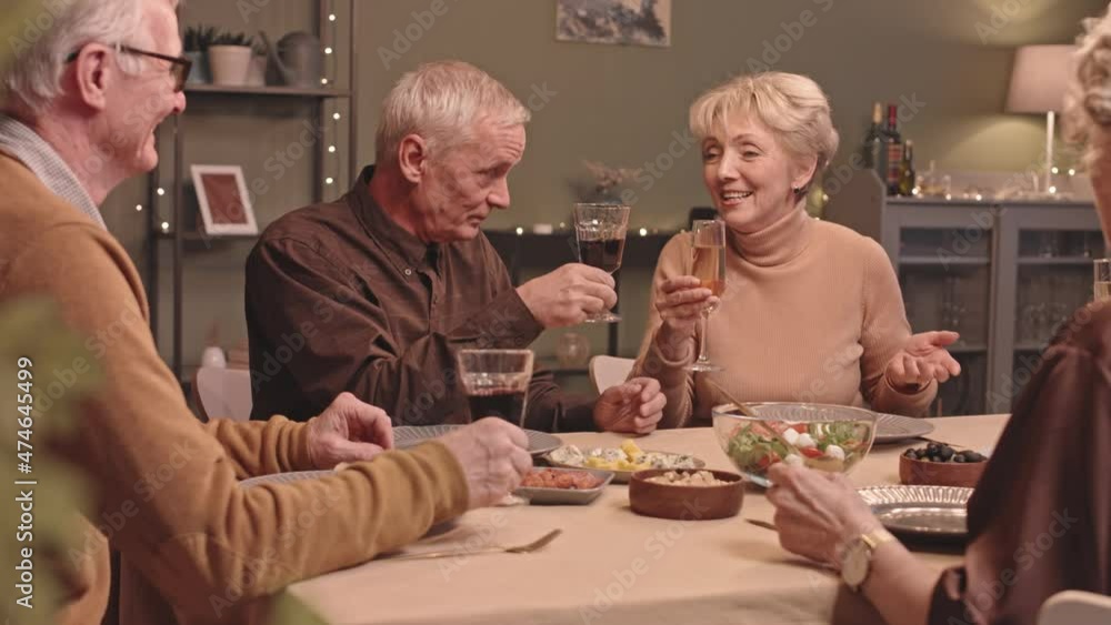 Waist up slowmo shot of senior friends sitting at festive dinner table at home, having conversation and toasting wine and champagne glasses, spending good time together Waist up slowmo shot of senior friends sitting at festive dinner table at home, having conversation and toasting wine and champagne glasses, spending good time together