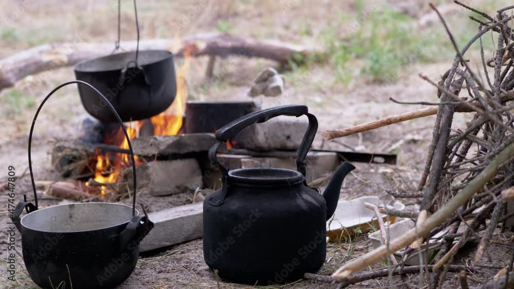 Kettle and Saucepan on Against the Background of Preparing Food on a Campfire. A male tourist puts a boiling cauldron of water on the grate of the fire. Concept of survival, trip, hiking, camping. 4K, Kettle and Saucepan on Against the Background of Preparing Food on a Campfire. A male tourist puts a boiling cauldron of water on the grate of the fire. Concept of survival, trip, hiking, camping. 4K,
