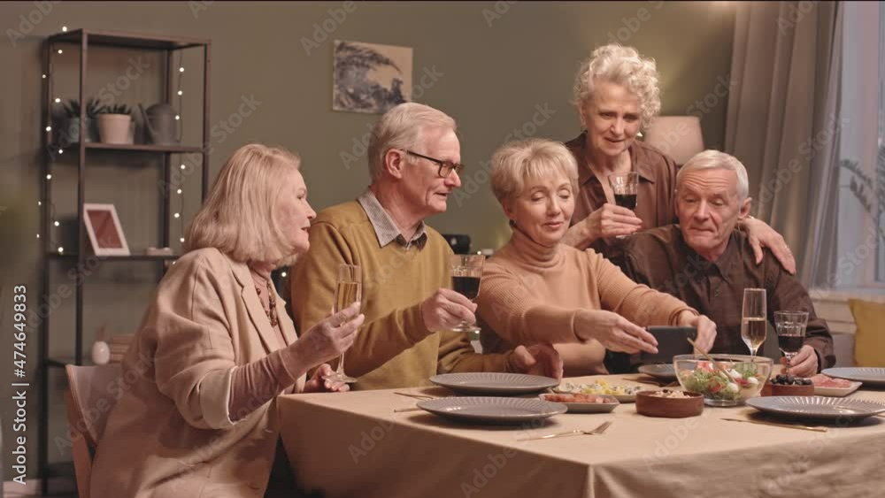 Waist up slowmo shot of five cheerful senior men and women video chatting on smartphone with their friend or relative while having home dinner party together clinking glasses at festive table Waist up slowmo shot of five cheerful senior men and women video chatting on smartphone with their friend or relative while having home dinner party together clinking glasses at festive table