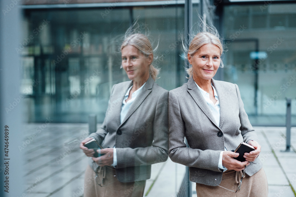 Smiling businesswoman with mobile phone leaning on glass wall Smiling businesswoman with mobile phone leaning on glass wall