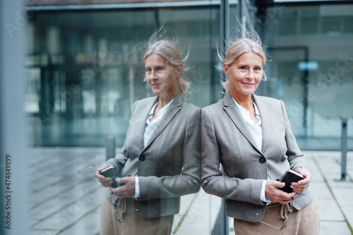 Smiling businesswoman with mobile phone leaning on glass wall