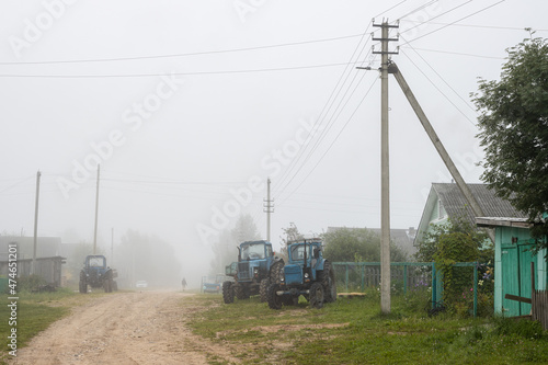 Misty morning in the countryside. View of the village street. Old agricultural tractors on the side of a dirt road. Lonely man in the fog. Foggy rural landscape. Everyday village life. Russia.