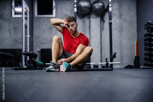 Exhaustion in training. The man sits on the floor of the gym and rests between exercises. He is tired and wipes the sweat from his face with his hand. Persistence and weight loss