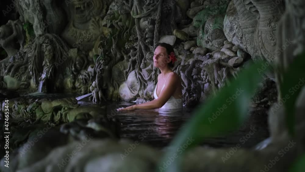 Beautiful woman with red flower in hair meditating and bathing in Bali water pond with Indonesian sculptures in tropics. Beautiful woman with red flower in hair meditating and bathing in Bali water pond with Indonesian sculptures in tropics.
