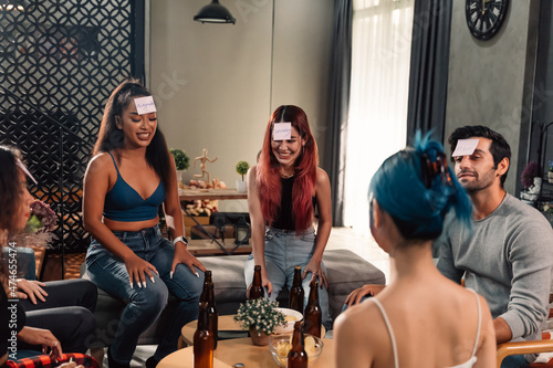 Group of male and female happy friends drinking beer and playing game guess who while having fun at home during housewarming party and reunion