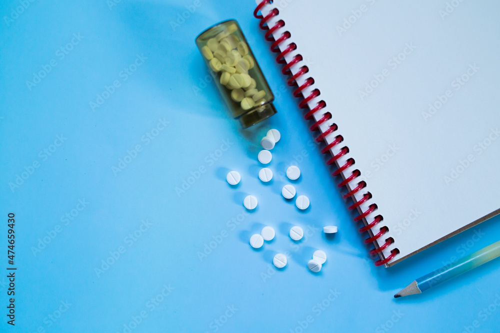 White medical pills and tablets spilling out of a drug bottle on a blue background. Medical layout pill, notebook and pencil copy space. White medical pills and tablets spilling out of a drug bottle on a blue background. Medical layout pill, notebook and pencil copy space.