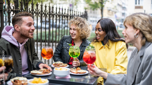 Multiracial happy young people eating and drinking, diverse cheerful mates laughing enjoying meal, having fun sitting together at restaurant table