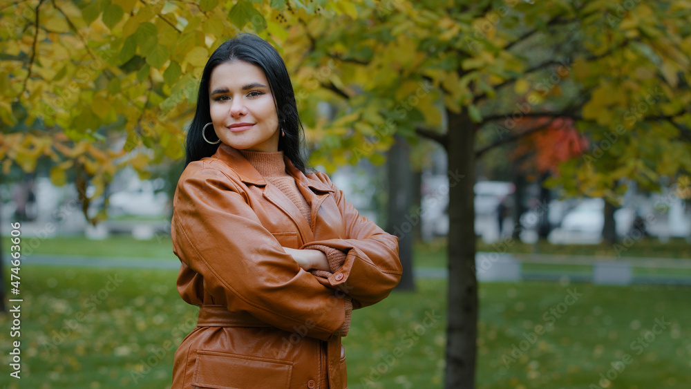 Beautiful young elegant woman looking into distance crossed arms over chest turns slowly focuses gaze on camera smiles shyly posing attractive pretty Hispanic girl stand in city autumn park outdoors Beautiful young elegant woman looking into distance crossed arms over chest turns slowly focuses gaze on camera smiles shyly posing attractive pretty Hispanic girl stand in city autumn park outdoors