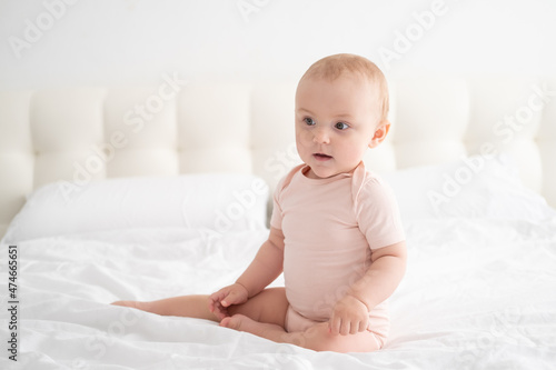 portrait of baby girl 10 months in light pink bodysuit on white bedding on bed