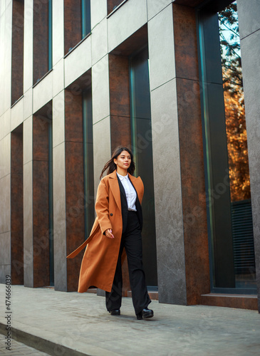 Asian business woman in long beige coat and hat walks down modern street on background of building in urban style © okostia