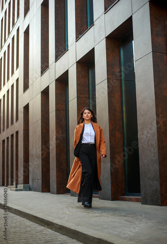 Asian business woman in long beige coat and hat walks down modern street on background of building in urban style
