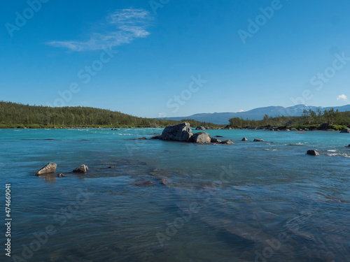 View of turquoise blue water of wild Vuojatadno river. Northern landscape in Swedish Lapland with birch tree forest and green mountains at Padjelantaleden hiking trail. Summer sunny day, blue sky photo