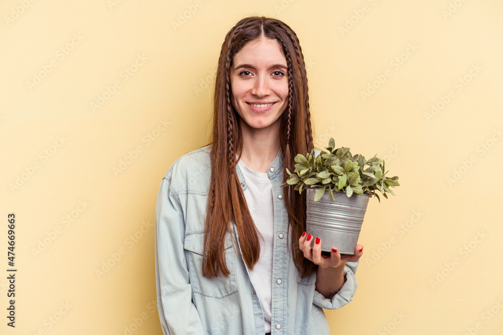 Young caucasian woman holding a plant isolated on yellow background happy, smiling and cheerful. Young caucasian woman holding a plant isolated on yellow background happy, smiling and cheerful.