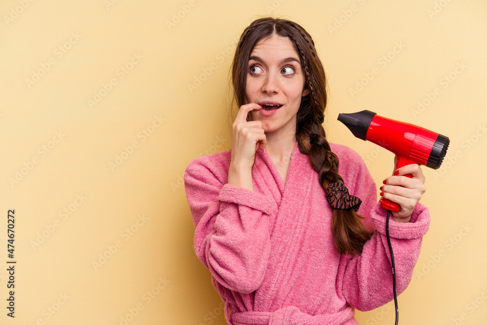 Young woman holding a hairdryer isolated on yellow background relaxed thinking about something looking at a copy space. Young woman holding a hairdryer isolated on yellow background relaxed thinking about something looking at a copy space.