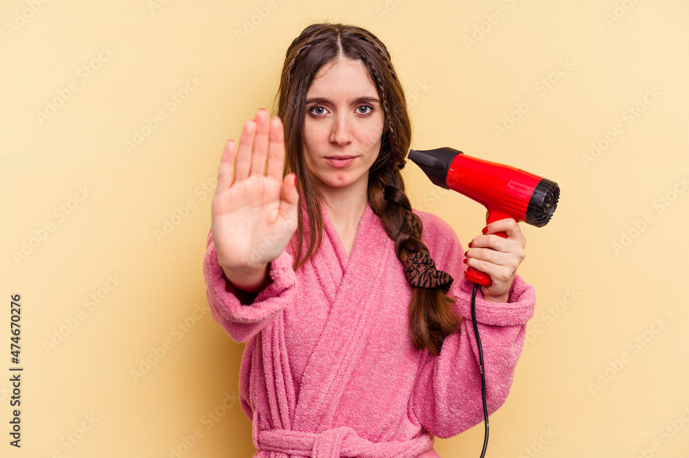 Young woman holding a hairdryer isolated on yellow background standing with outstretched hand showing stop sign, preventing you. Young woman holding a hairdryer isolated on yellow background standing with outstretched hand showing stop sign, preventing you.