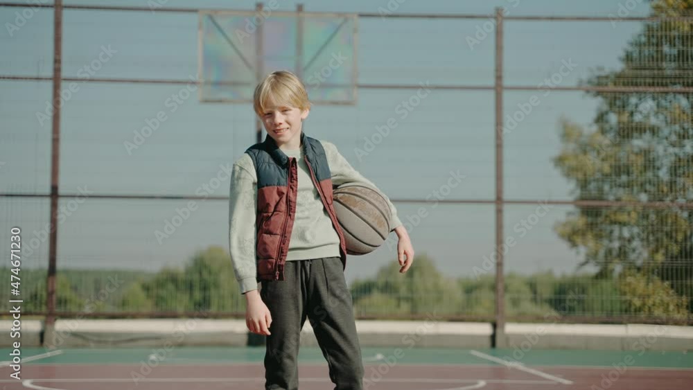 Boy stands on a court holding basketball ball and looking at the camera. Boy training street basketball. Focused streetball player practicing basketball alone outdoor. High quality 4k footage Boy stands on a court holding basketball ball and looking at the camera. Boy training street basketball. Focused streetball player practicing basketball alone outdoor. High quality 4k footage