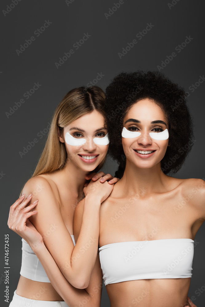joyful multiethnic women in hydrogel eye patches smiling at camera isolated on grey joyful multiethnic women in hydrogel eye patches smiling at camera isolated on grey