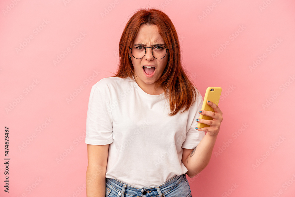 Young caucasian woman holding a mobile phone isolated on pink background screaming very angry and aggressive. Young caucasian woman holding a mobile phone isolated on pink background screaming very angry and aggressive.