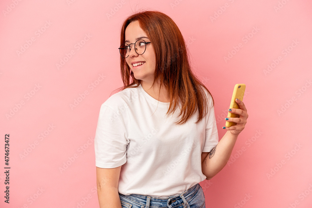Young caucasian woman holding a mobile phone isolated on pink background looks aside smiling, cheerful and pleasant. Young caucasian woman holding a mobile phone isolated on pink background looks aside smiling, cheerful and pleasant.