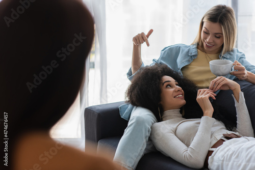 cheerful woman with cup of tea pointing at african american girlfriend near blurred psychologist