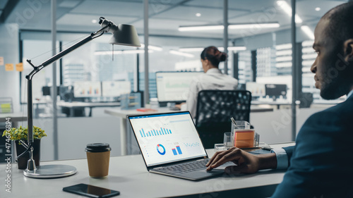 Successful Black Businessman Sitting at Desk Working on Laptop Computer in Big City Office. Hard Working Top Manager Doing Research for e-Commerce Project with Graps, Charts and Tables. Over Shoulder