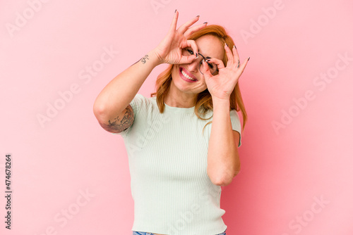 Young caucasian woman isolated on pink background showing okay sign over eyes © Asier