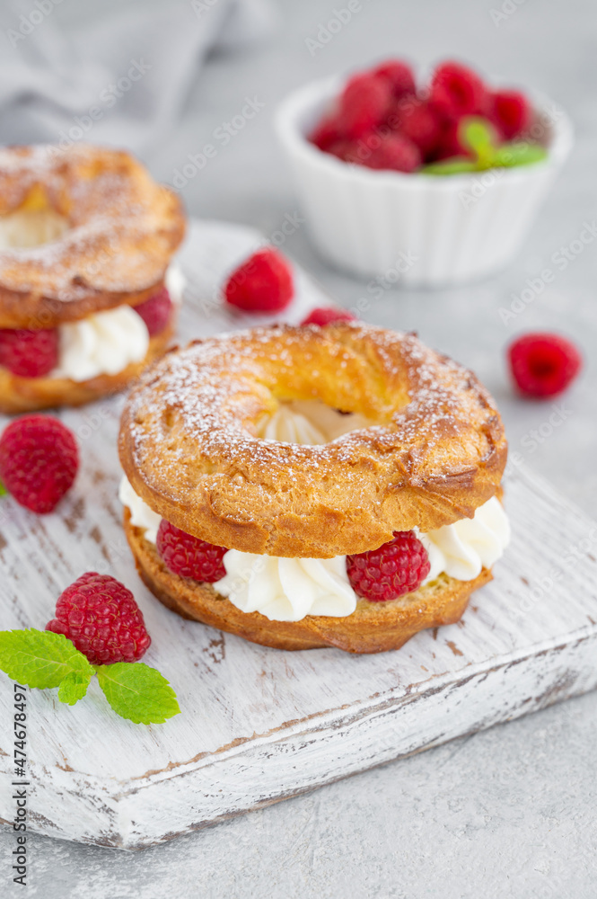 Choux pastries. Choux rings with cream of cream cheese or cottage cheese and fresh raspberries, dusted with powdered sugar on a wooden board on a gray concrete background.