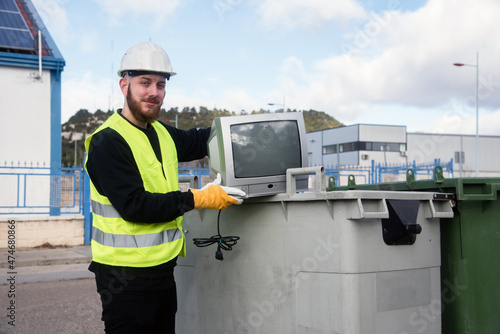 Trabajador de la planta de reciclaje posando junto a un viejo televisor y un contenedor de basura. Hombre joven con chaleco reflectante y guantes y casco protector photo