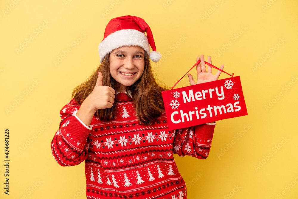 Little caucasian girl holding ‘’merry Christmas’’ placard isolated on yellow background smiling and raising thumb up Little caucasian girl holding ‘’merry Christmas’’ placard isolated on yellow background smiling and raising thumb up