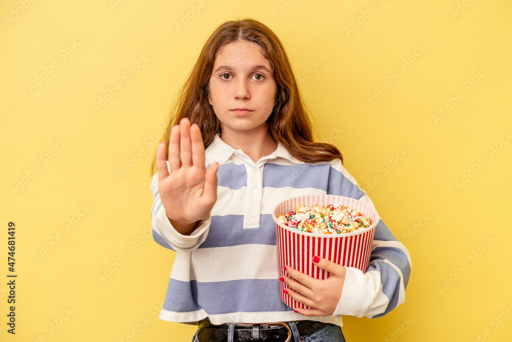 Little caucasian girl holding popcorns isolated on yellow background standing with outstretched hand showing stop sign, preventing you. Little caucasian girl holding popcorns isolated on yellow background standing with outstretched hand showing stop sign, preventing you.