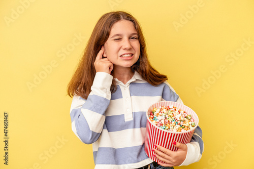 Little caucasian girl holding popcorns isolated on yellow background covering ears with hands.