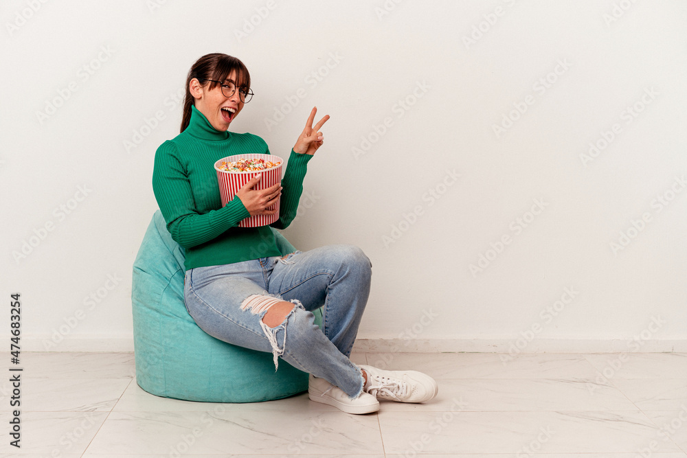 Young Argentinian woman eating popcorns on a puff isolated on white background joyful and carefree showing a peace symbol with fingers. Young Argentinian woman eating popcorns on a puff isolated on white background joyful and carefree showing a peace symbol with fingers.