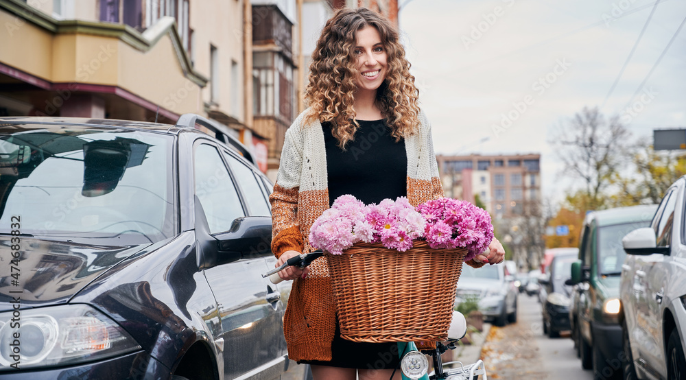 Beautiful young smiling girl with curly hair standing in the middle of city street with traffic jam, holding bicycle handlebar with full flowers wicker basket.